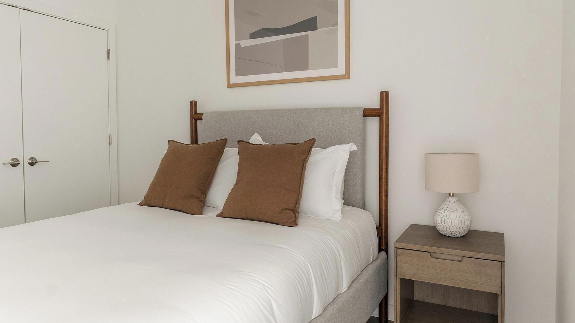 A neatly made bed with a headboard, a bedside table with a lamp, and a door to the left.
