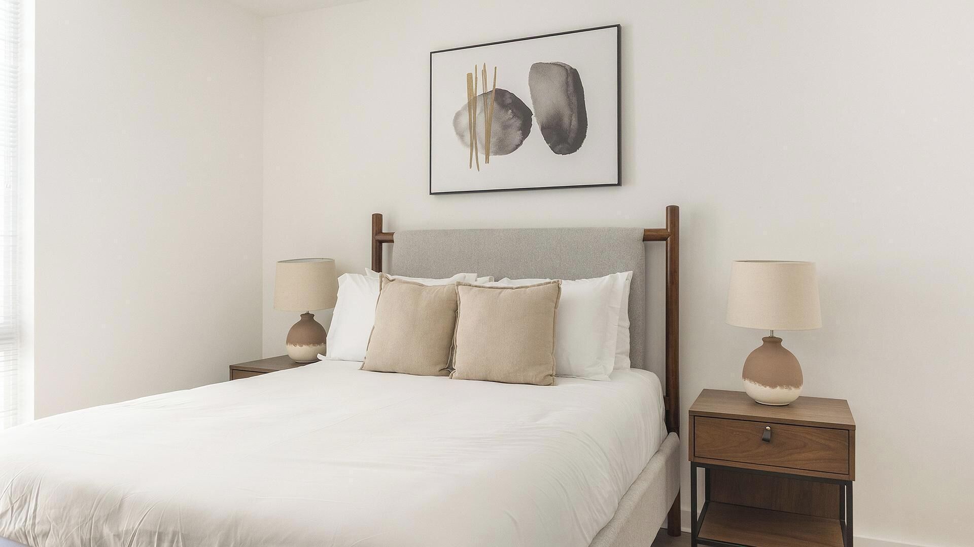 A neatly made bed with a headboard, two bedside tables with lamps, and a wall-mounted abstract painting.