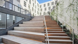 A staircase with wooden steps and metal railings, leading to a modern building with large windows and a concrete wall with climbing plants. - Landing Modern 1bd, Gym, Yoga Studio (Durham)