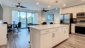 Una cocina moderna con gabinetes blancos, una isla central y electrodomésticos integrados. Cuenta con un comedor con mesa y sillas, y un ventilador de techo.