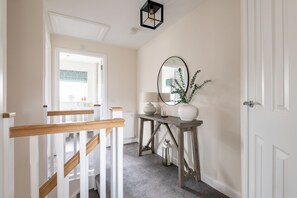 A hallway with a wooden railing, a table with decorative items, a mirror, and a door. - Annie Paton Road (No 123) | St Andrews (St Andrews)