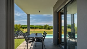 Eine moderne Terrassengestaltung mit Tisch und Stühlen, eine Glastür, die zu einem Pool führt, und Blick auf eine grüne Landschaft.