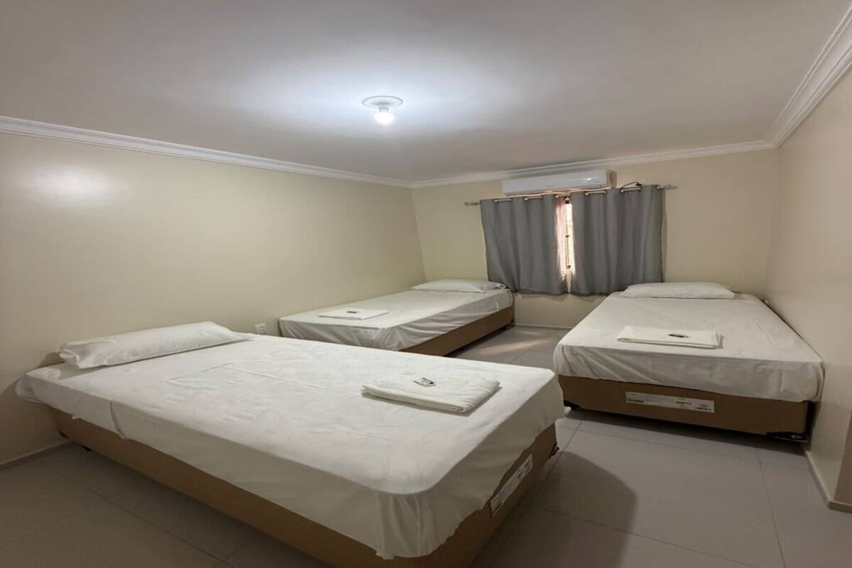 A room with three single beds, a ceiling light, an air conditioning unit, and a window with curtains.