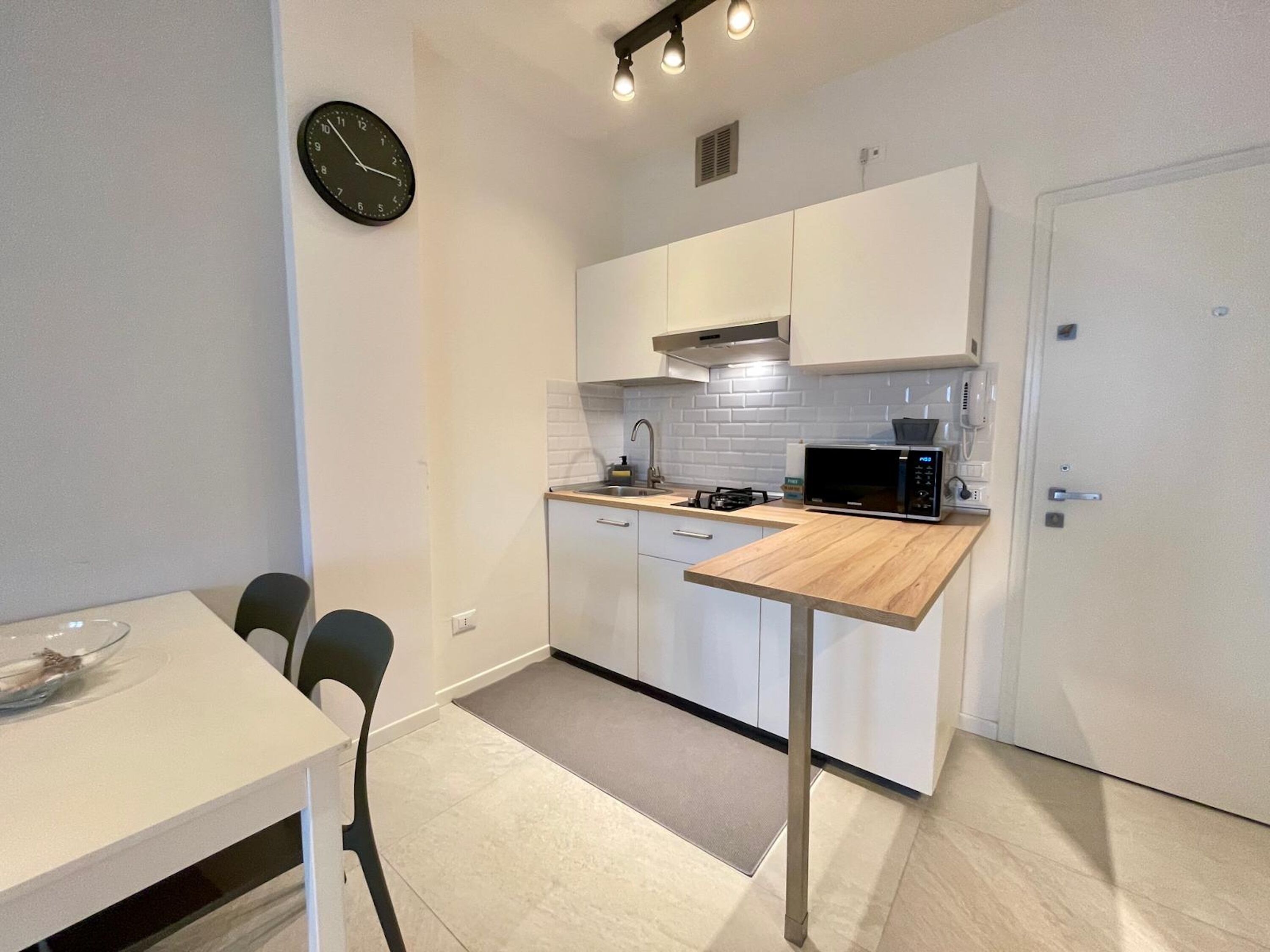 Une cuisine compacte avec des meubles blancs, un four à micro-ondes, un évier et une horloge au mur.