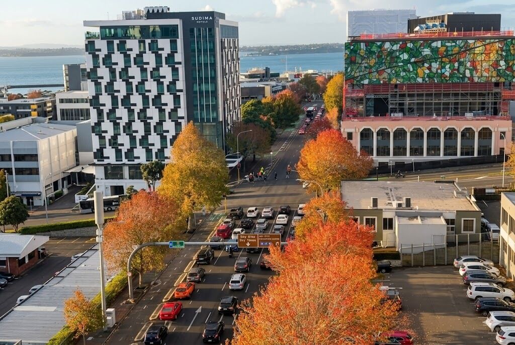 Een modern gebouw met een unieke geometrische gevel, een parkeerplaats met auto's en herfstbomen.