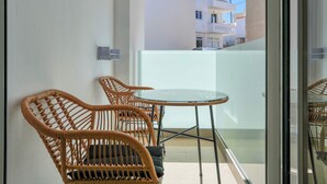 Uma varanda com cadeiras de vime, uma mesa de vidro, uma parede branca e vista para edifícios ao fundo.