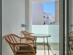 Ein Balkon mit Korbstühlen und einem Glastisch, eine weiße Wand und im Hintergrund ein Blick auf Gebäude.