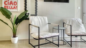 Uma sala de estar moderna com dois poltronas, uma mesinha redonda, uma planta em vaso, um espelho e uma TV de tela plana na parede.