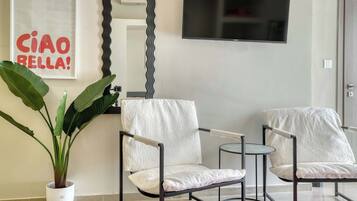 A modern living room with two armchairs, a small round table, a potted plant, a mirror, and a flat-screen TV mounted on the wall.