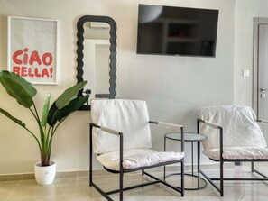 Ein modernes Wohnzimmer mit zwei Sesseln, einem kleinen runden Tisch, einer Topfpflanze, einem Spiegel und einem an der Wand befestigten Flachbildfernseher.