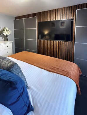 Ein modernes Schlafzimmer mit einem Bett, einem an einer Holzwand montierten Fernseher, einer weißen Kommode und einer Vase mit Blumen.