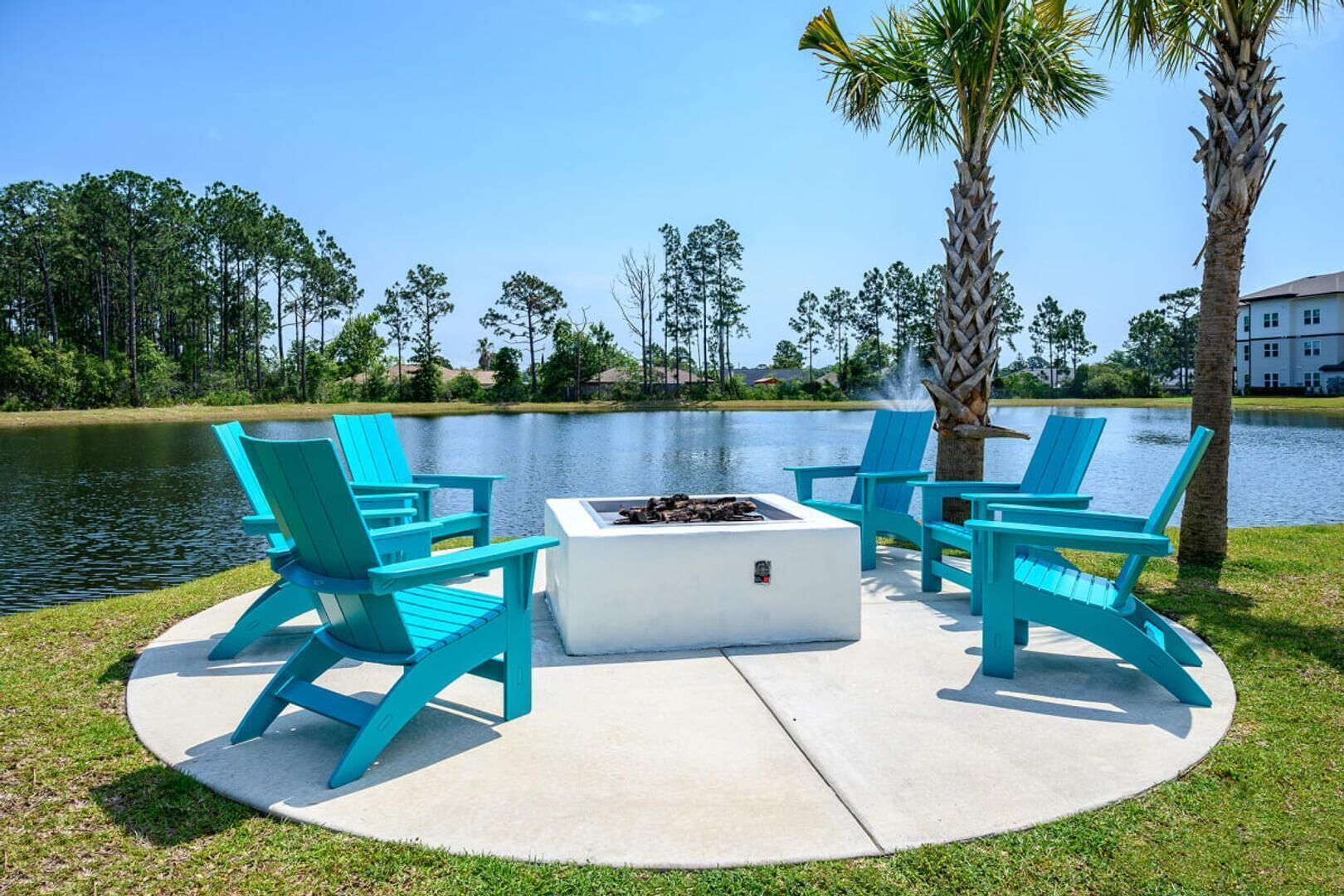 A circular outdoor seating area with a fire pit, surrounded by turquoise chairs, near a body of water with trees and a building in the background.