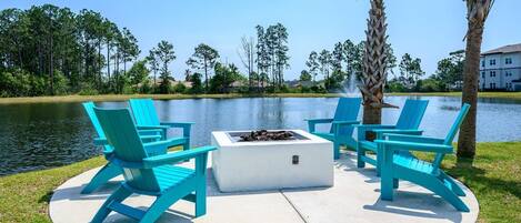 A circular outdoor seating area with a fire pit, surrounded by turquoise chairs, near a body of water with trees and a building in the background.
