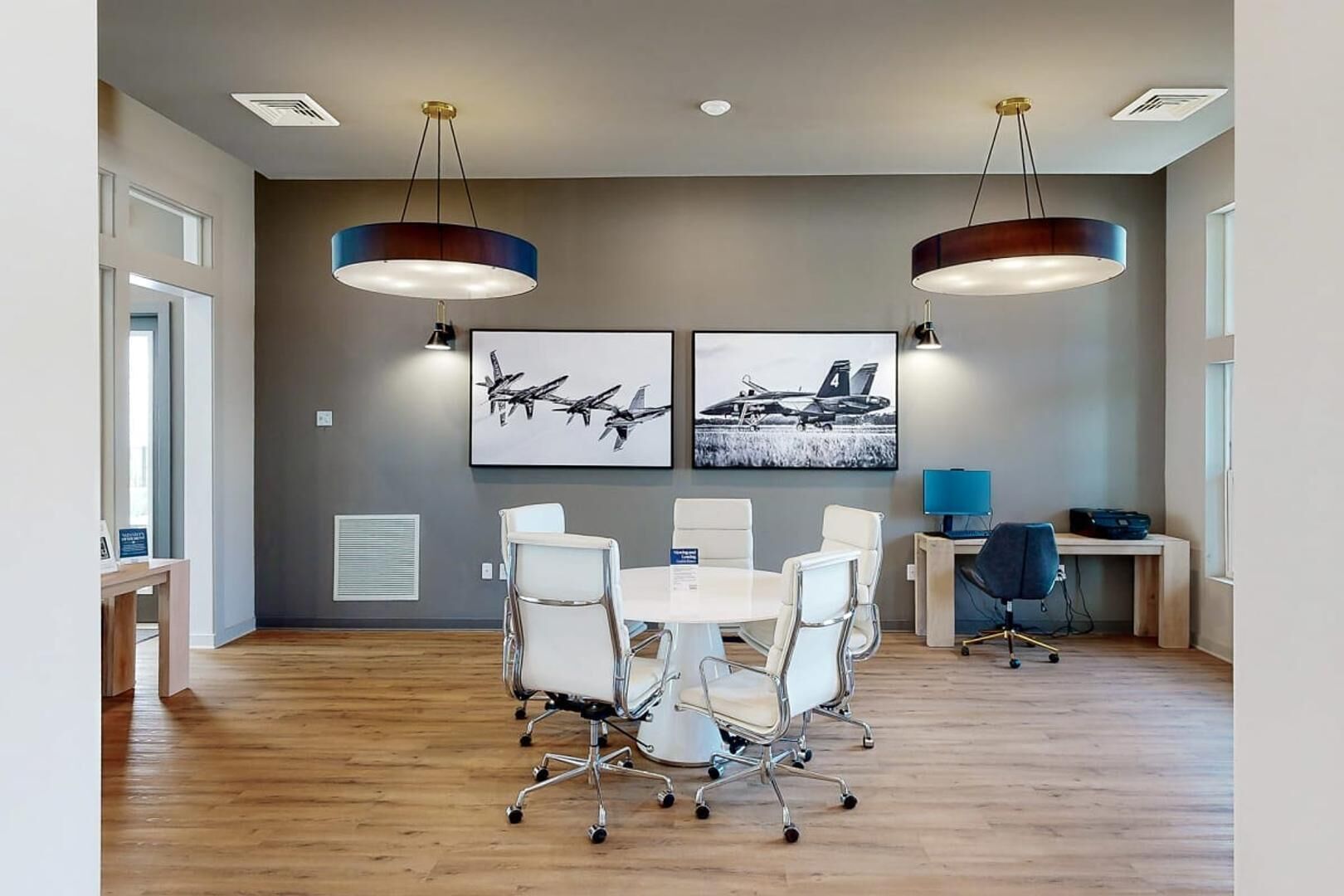 Une salle de conférence avec une table ronde, des chaises blanches, deux grands luminaires suspendus et deux tableaux encadrés au mur.