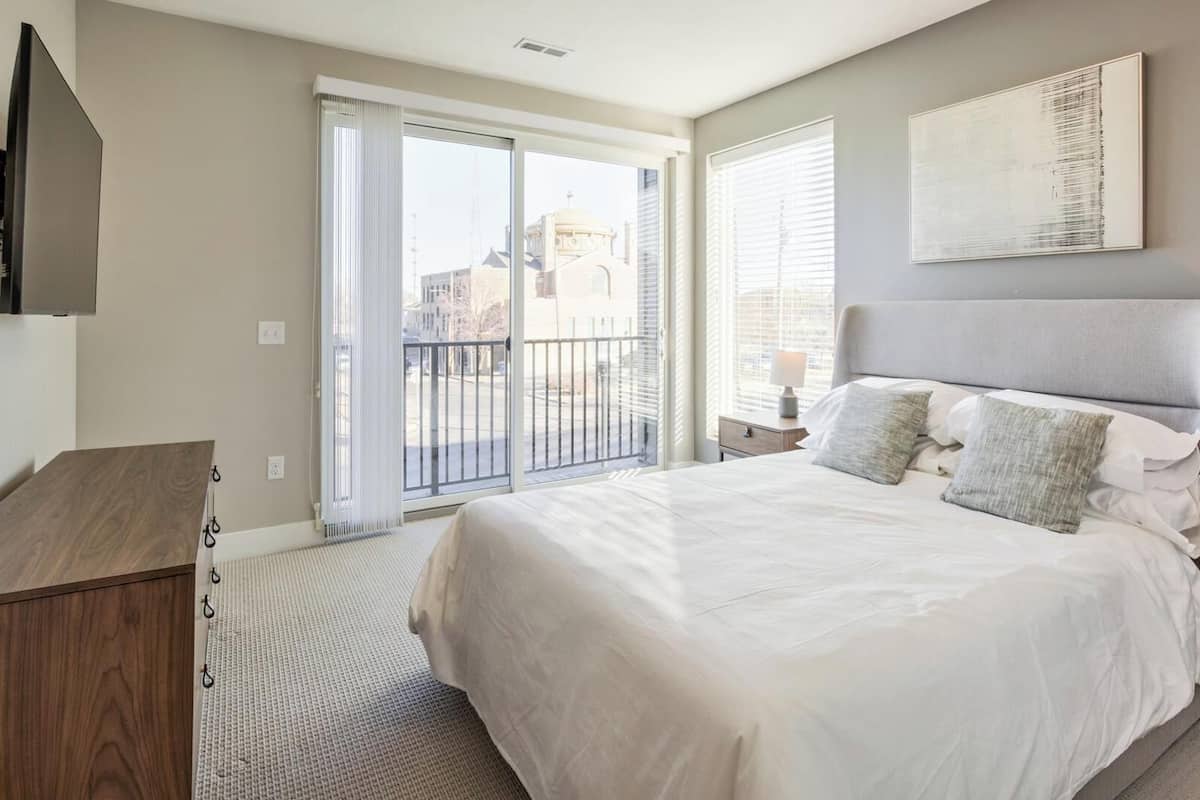 A bedroom with a bed, a TV mounted on the wall, a window with blinds, and a view of the outside.