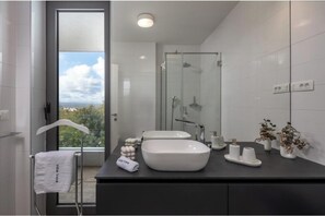 Un bagno moderno con una grande finestra, un box doccia in vetro e un piano d'appoggio scuro con due lavabi e oggetti decorativi.