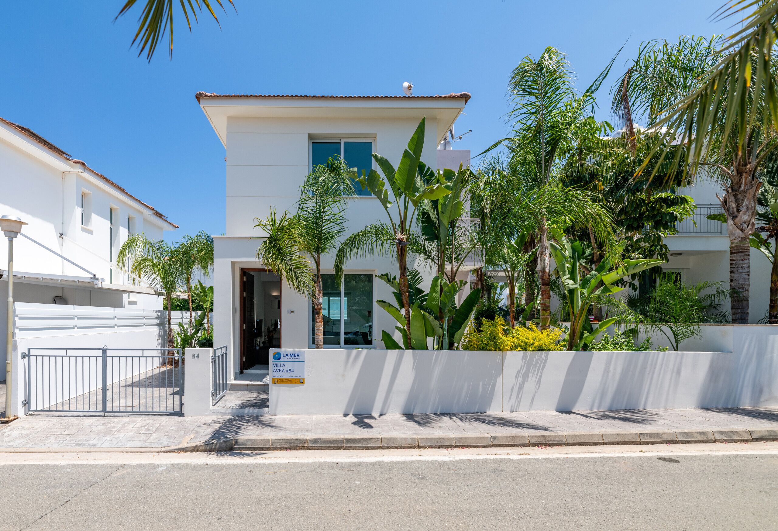 A modern building with a white facade, a gated entrance, and lush tropical plants.