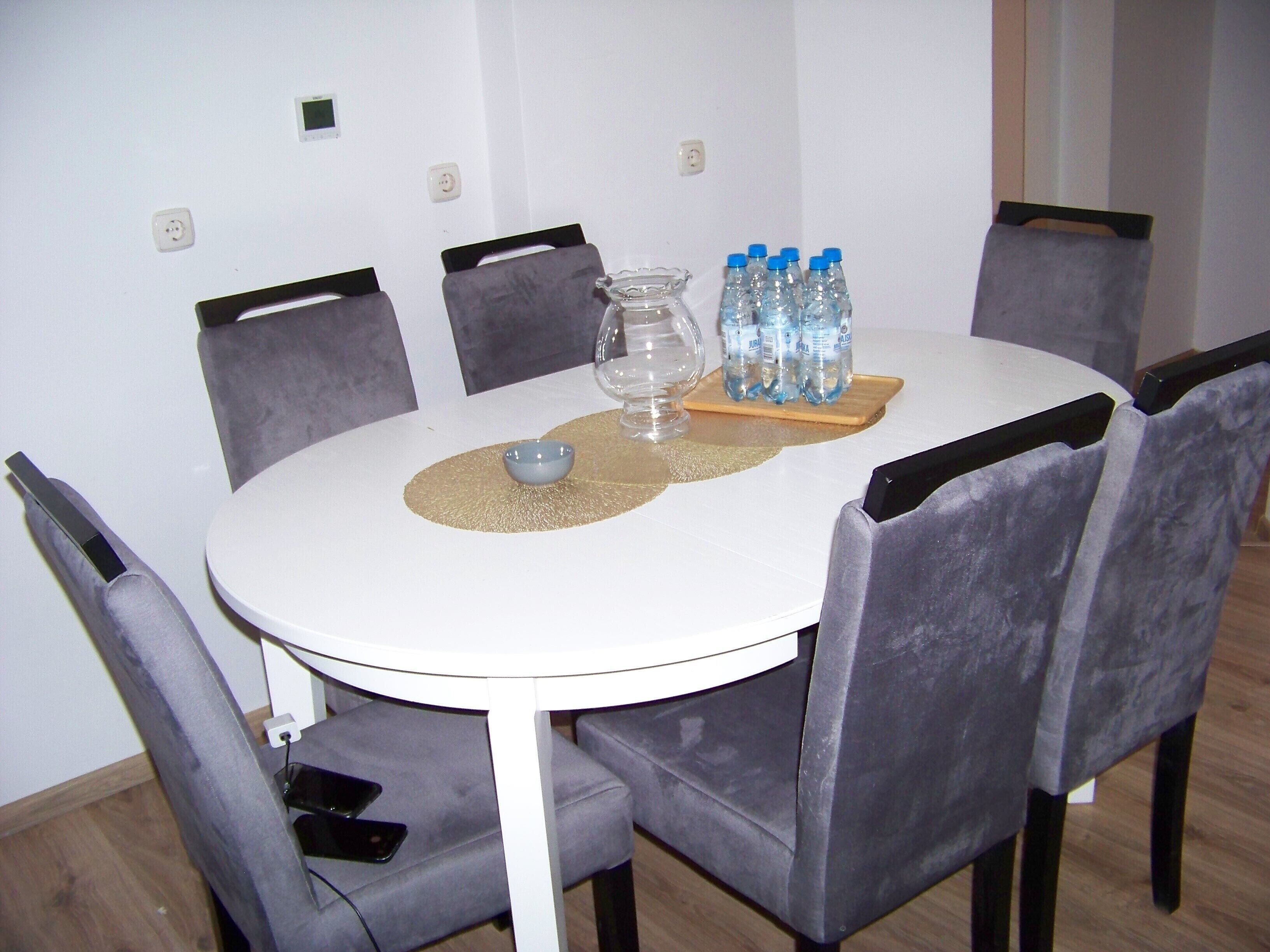 Ein runder Esstisch mit weißer Tischplatte, umgeben von sechs grauen, gepolsterten Stühlen. Auf dem Tisch steht eine transparente Glasvase, eine kleine blaue Schale und ein Holzbrett mit mehreren Wasserflaschen.