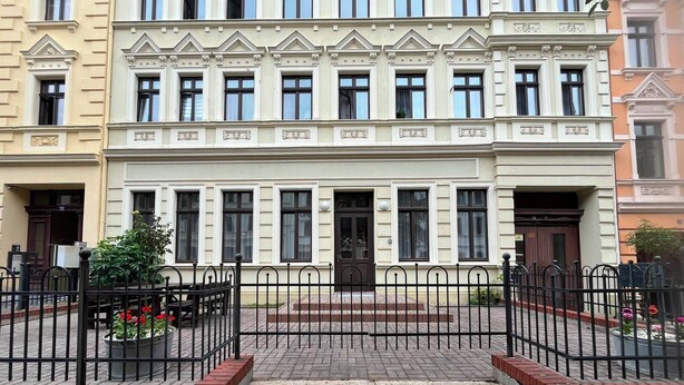 Ein mehrgeschossiges Gebäude mit einer symmetrischen Fassade, großen Fenstern und einem zentralen Eingang mit Stufen.