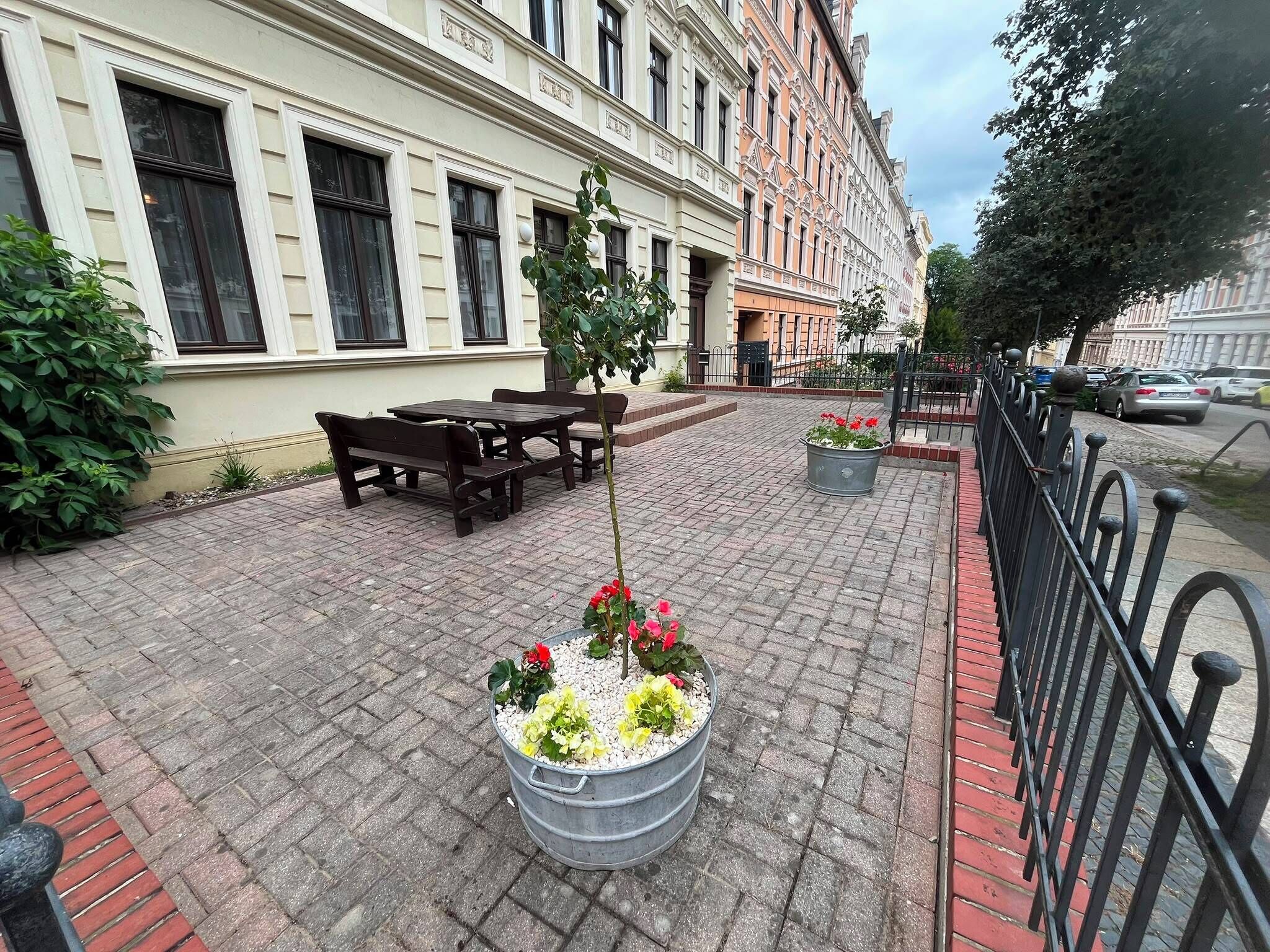 A paved outdoor area with tables and benches, a metal planter with flowers, and a building with classical architecture.
