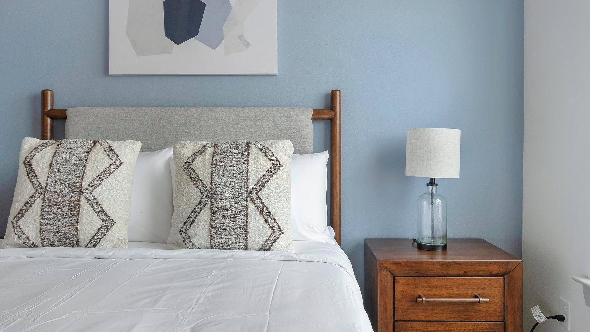 A bed with a wooden headboard, two patterned pillows, a white bedspread, a bedside table with a lamp, and a wall-mounted abstract artwork.