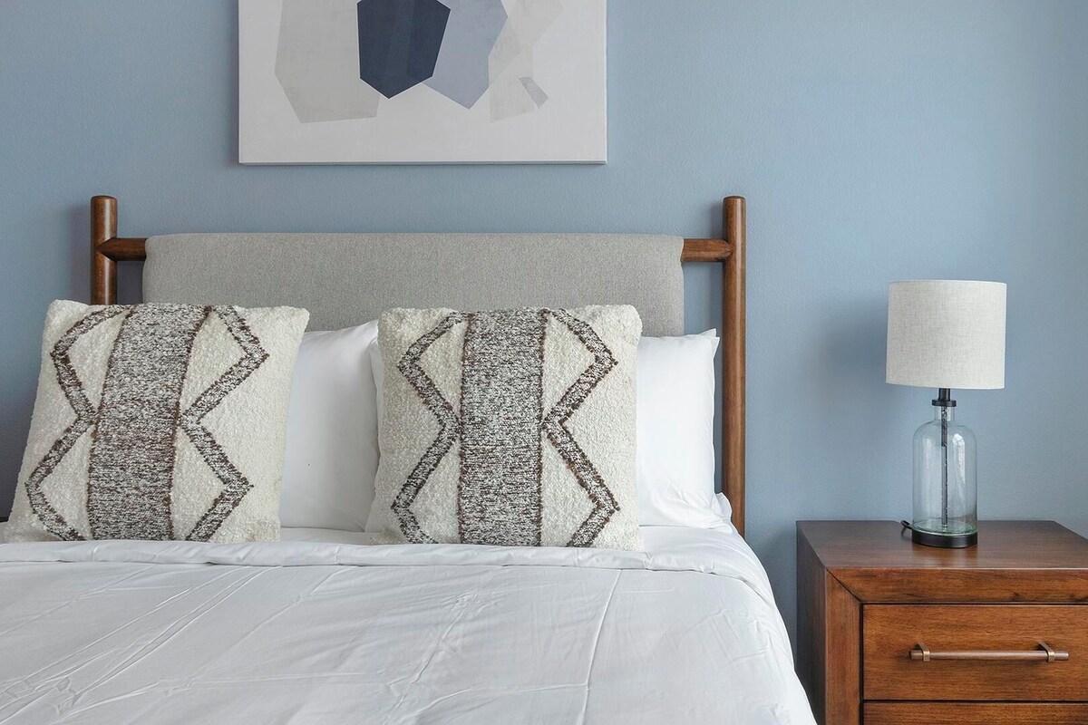 A bed with a wooden headboard, two patterned pillows, a white bedspread, a bedside table with a lamp, and a wall-mounted abstract artwork.