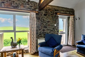 Ein Zimmer mit Steinwand, einem Holztisch mit einer Hahn-Dekoration, einem blauen Sessel und Blick auf grüne Felder durch ein Fenster.