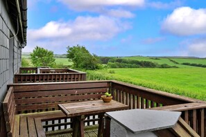 A wooden deck with a table and bench, overlooking a green field and hills. - Mucklow, Quinceborough Farm (Widemouth Bay)