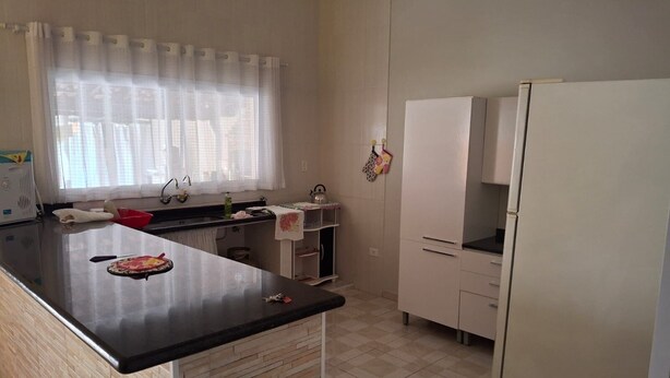 A kitchen with a black countertop, white cabinets, a refrigerator, and a window with a white curtain.