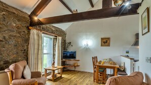 A cozy living room with a stone wall, wooden furniture, a television, and a dining area. - Squire, Quinceborough Farm (Widemouth Bay)