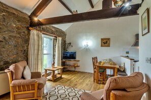 A cozy living room with a stone wall, wooden furniture, a television, and a dining area. - Squire, Quinceborough Farm (Widemouth Bay)