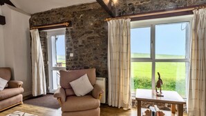 A room with a stone wall, a wooden ceiling, a sofa, a chair, a table with books, and a view of greenery outside. - Squire, Quinceborough Farm (Widemouth Bay)