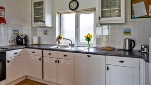 A kitchen with white cabinets, a black countertop, a window with a view, a clock, and a corkboard with papers. - Squire, Quinceborough Farm (Widemouth Bay)