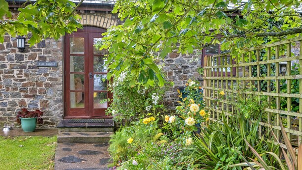 A stone building with a wooden door, a garden with various plants and flowers, and a wooden fence. - Roost, Quinceborough Farm (Widemouth Bay)