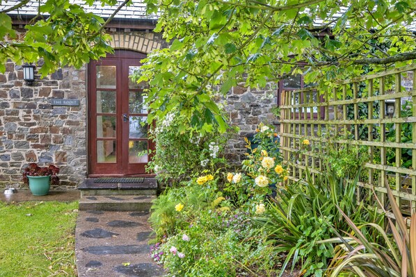 A stone building with a wooden door, a garden with various plants and flowers, and a wooden fence. - Roost, Quinceborough Farm (Widemouth Bay)