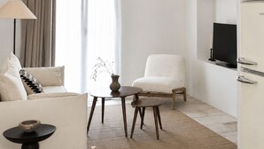 Una sala de estar moderna con sofá, sillón, mesa de comedor con un tazón de alcachofas, lámpara y televisor en la pared.