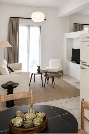 A modern living room with a sofa, armchair, dining table with a bowl of artichokes, a lamp, and a television on a wall. - Lanu Sea View Studio (Argostoli)
