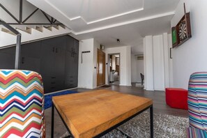 Un salón moderno con una mesa de centro de madera, sillas coloridas, un dardo y una escalera con un diseño geométrico en el techo.