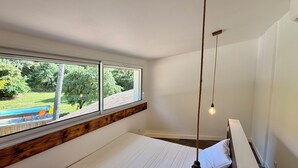 Quarto com uma janela ampla, cama com roupa de cama branca, luminária pendente e vista para uma piscina e vegetação lá fora.