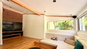 Uma sala de estar moderna com piso de madeira, uma TV de tela plana, um sofá, uma janela com vista para vegetação e luminárias embutidas no teto.