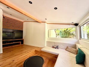 A modern living room with a wooden floor, a flat-screen TV, a sofa, a window with a view of greenery, and recessed ceiling lights. - Vacation Villa (Méounes-lès-Montrieux)