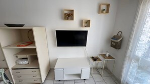 Huoneessa on seinälle asennettu televisio, laatikoilla varustettu työpöytä, hylly, jossa on tavaroita, ja pieni pöytä, jonka päällä on kori.