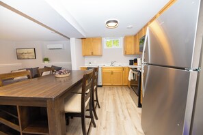 A modern kitchen with wooden cabinets, a stainless steel refrigerator, a dining table with chairs, and a window with a view of greenery.