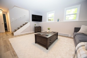 A living room with a staircase, a flat-screen TV mounted on the wall, and a window with a view of trees.