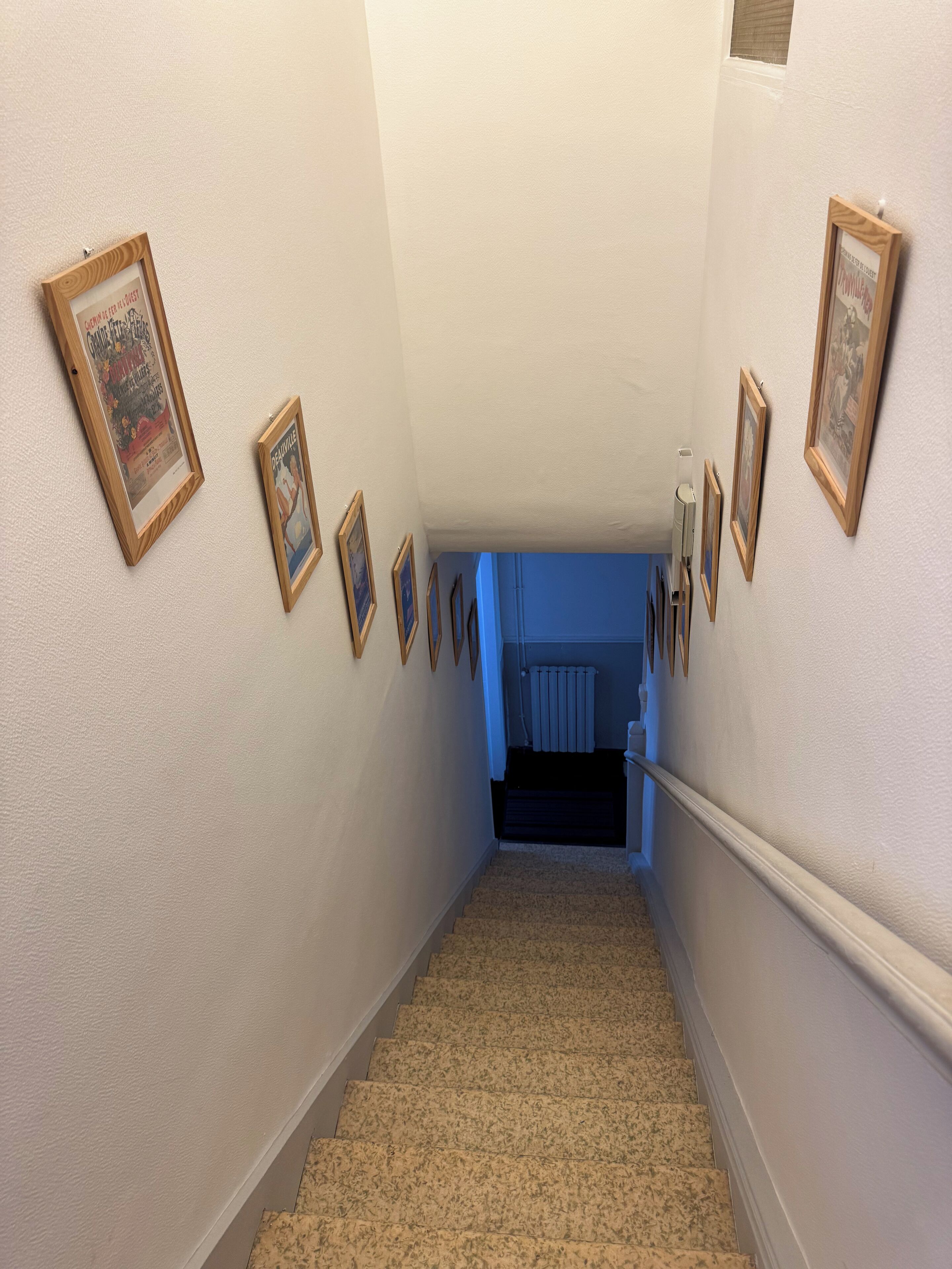 Un escalier recouvert de moquette, orné de tableaux encadrés le long des murs, mène à un autre escalier doté d’une rampe bleue.