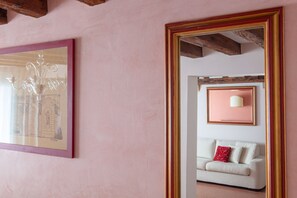 Een kamer met een grote spiegel, een ingelijst kunstwerk, een witte bank met een rood kussen, en een lamp aan de muur.