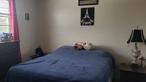 Une chambre à coucher avec un lit, une commode, une fenêtre avec des rideaux, une lampe et un tableau représentant la Tour Eiffel.