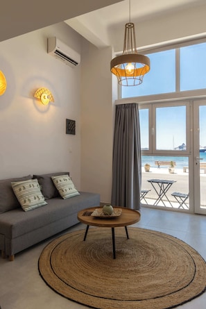 A living room with a sofa, a round coffee table, a hanging light fixture, and a view of the sea through large windows. - Akti Waterfront Modern lofts: Aegean (Adamantas Milos)