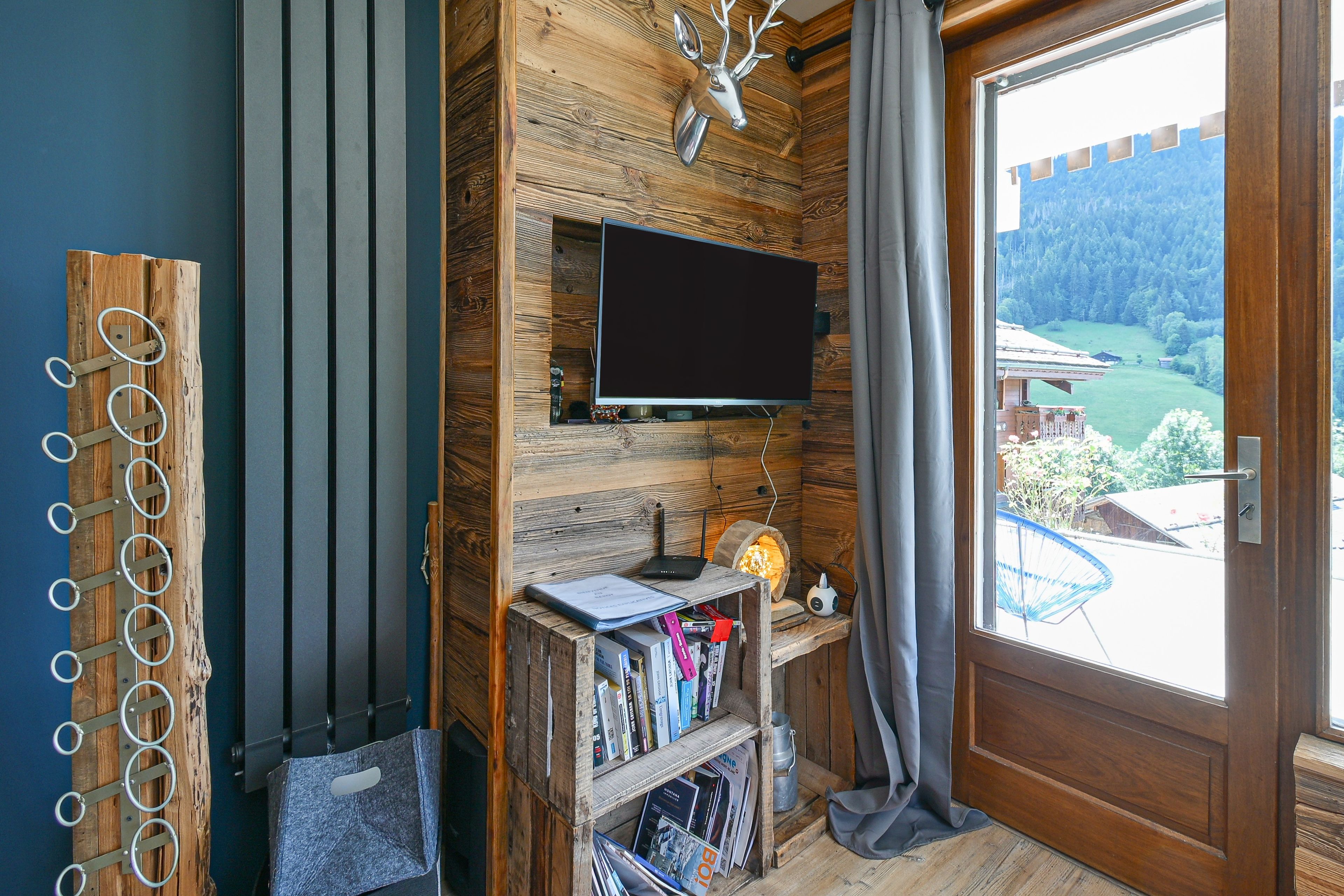 Une pièce dotée d’un téléviseur fixé au mur, de lambris en bois, d’une bibliothèque remplie de livres et offrant une vue sur un paysage verdoyant par une porte.
