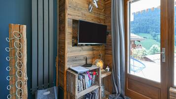 Une pièce dotée d’un téléviseur fixé au mur, de lambris en bois, d’une bibliothèque remplie de livres et offrant une vue sur un paysage verdoyant par une porte.